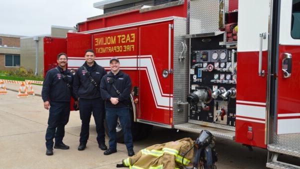 Three fire fighters stand in front of a red fire truck outside near a pile of protective gear.